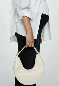 Hobo bag in soft cream faux leather with a curved silhouette, featuring a top handle with knotted accents. Worn on the arm, paired with black pants.