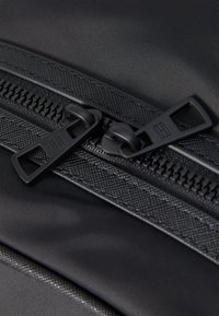 Black, textured fabric bag featuring a smooth zipper with two black pull tabs, highlighted by a subtle sheen and detailed stitching.