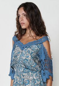Plava haljina s paisley uzorkom, s čipkastim detaljima na ramenima, koja sadrži cvjetne akcentne i blago teksturiranu tkaninu.