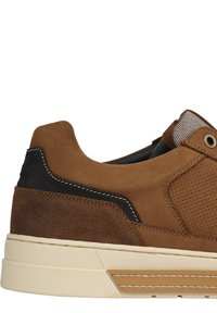 Brown suede sneaker with perforated detailing, black accents, and a cream rubber sole. Features a rounded toe and a padded collar.