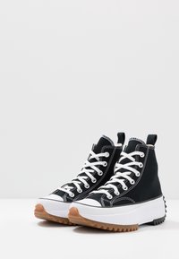 Czarne sneakersy high-top na płaskiej podeszwie z białymi sznurówkami, masywną białą i brązową podeszwą oraz teksturowaną gumową wkładką dla lepszej przyczepności.
