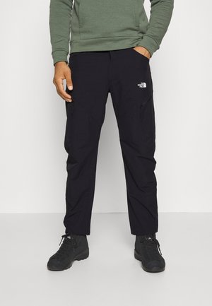 Hombre con pantalones negros de North Face con bolsillos con cremallera, sudadera verde y botas de senderismo negras, de pie en el interior sobre un suelo claro.