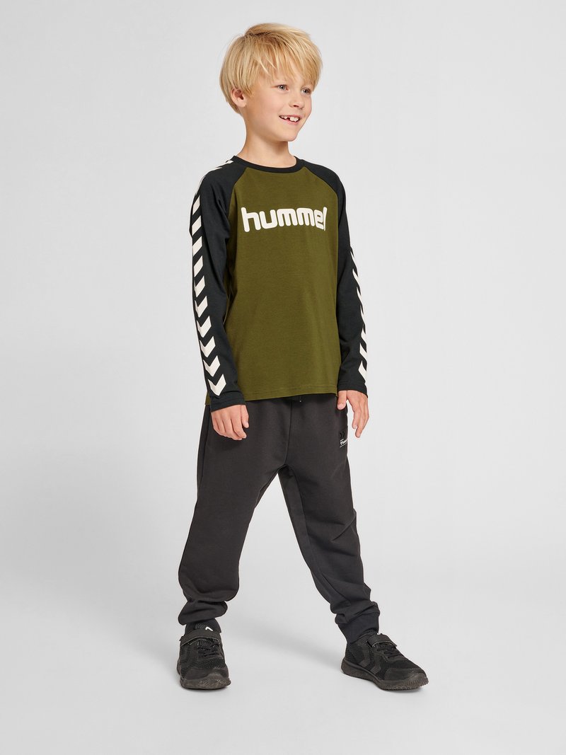 Blond jongetje staat, glimlacht, draagt een groen-zwarte lange mouwen shirt met de tekst "hummel" en zwarte broek en sneakers op een lichtgrijze achtergrond.