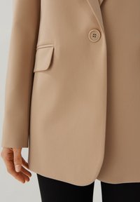 Detailní záběr na béžový blazer s předním knoflíkem, klopovou kapsou a viditelnou rukou po straně, nošený přes černé kalhoty.