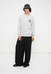 Sudadera gris con un estampado gráfico en el pecho, combinada con pantalones de pierna ancha a cuadros negros. Una gorra negra completa el conjunto.