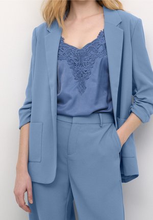 Kobieta nosząca jasnoniebieską marynarkę i pasujące spodnie z niebieską satynową koszulką wykończoną koronką, jedną ręką w kieszeni, a drugą swobodnie opadającą.