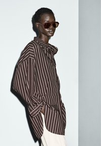 Bruin en wit gestreept oversized shirt met een puntige kraag, half geopende voorkant en opgerolde mouwen, gecombineerd met donkere zonnebril.