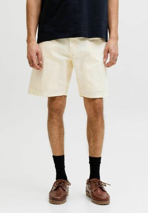 Mann trägt cremeweiße knielange Shorts, schwarze Socken, braune Lederschuhe und ein schwarzes T-Shirt, steht vor einem schlichten Hintergrund.