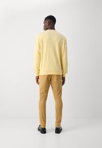 Gelbes Rundhals-Sweatshirt mit langen Ärmeln, kombiniert mit schmal geschnittenen hellbraunen Hosen und schwarzen Schuhen. Glatte Textur, lässige Passform.