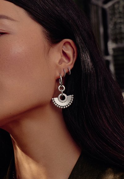 Το αριστερό αυτί μιας γυναίκας με τρία ασημένια κρίκους, συμπεριλαμβανομένου ενός μεγάλου με γεωμετρικό σχέδιο και μικρές ασημένιες χάντρες κατά μήκος του κάτω άκρου.