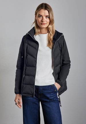 Giacca nera isolante con cappuccio, caratterizzata da un design trapuntato e accessori in argento. Indossata sopra un maglione bianco con texture e jeans blu.