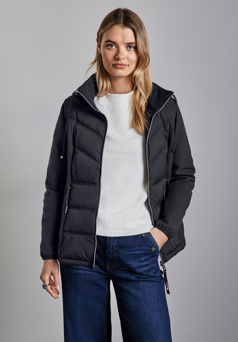 Svart isolerad jacka med huva, med en kviltad design och silverdetaljer. Buren över en vit texturerad tröja och blå jeans.
