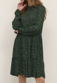 Vestido verde con estampado floral, mangas largas, escote redondo, cintura fruncida y falda acampanada. La tela presenta un delicado patrón de hojas.