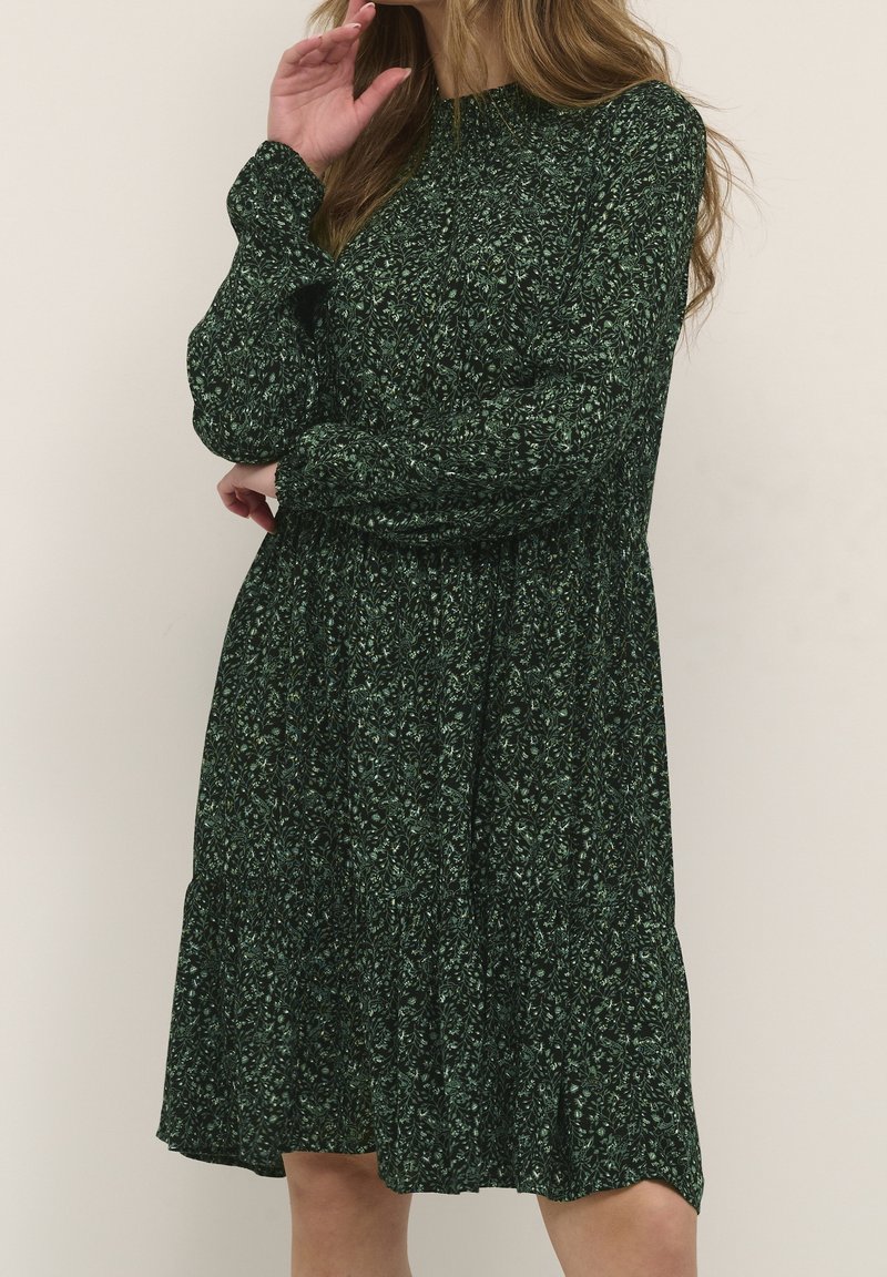 Vestido verde con estampado floral, mangas largas, escote redondo, cintura fruncida y falda acampanada. La tela presenta un delicado patrón de hojas.