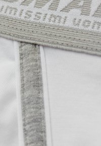 Weißes Kleidungsstück mit einem grauen strukturierten Taillenbund; es verfügt über ein gewebtes Logo und eine glatte Stoffstruktur mit Nähdetails nahe dem Taillenbund.