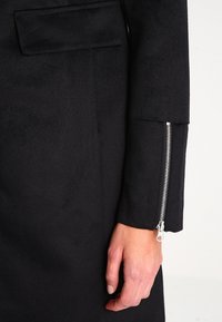 Tessuto di un cappotto nero con una texture liscia e dettagli con maniche con zip. Presenta una tasca verticale singola sulla parte superiore del braccio.