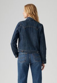 Tume sinine denimpusa, millel on sobiv disain, klassikaline krae ja terav yoke. Omab silmapaistvat õmblustööd ja metallnööpe.