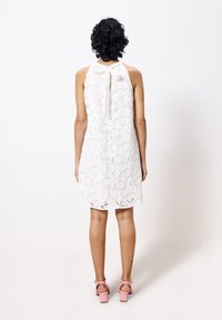 Weißes Spitzenkleid mit einer Schleifendetail am Hals, das ein Blumenmuster aufweist. Kombiniert mit rosa Absatzsandalen.