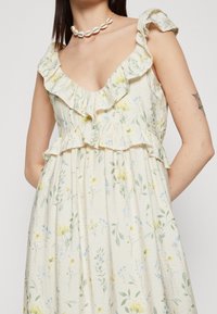Robe en coton à motifs floraux en crème avec un col et des bretelles à volants, présentant des motifs botaniques jaunes et bleus sur l'ensemble du tissu.