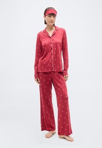 Femme debout, pieds nus, portant un pyjama boutonné à manches longues avec des motifs de cœurs rouges, un pantalon assorti et un masque de sommeil sur le front.