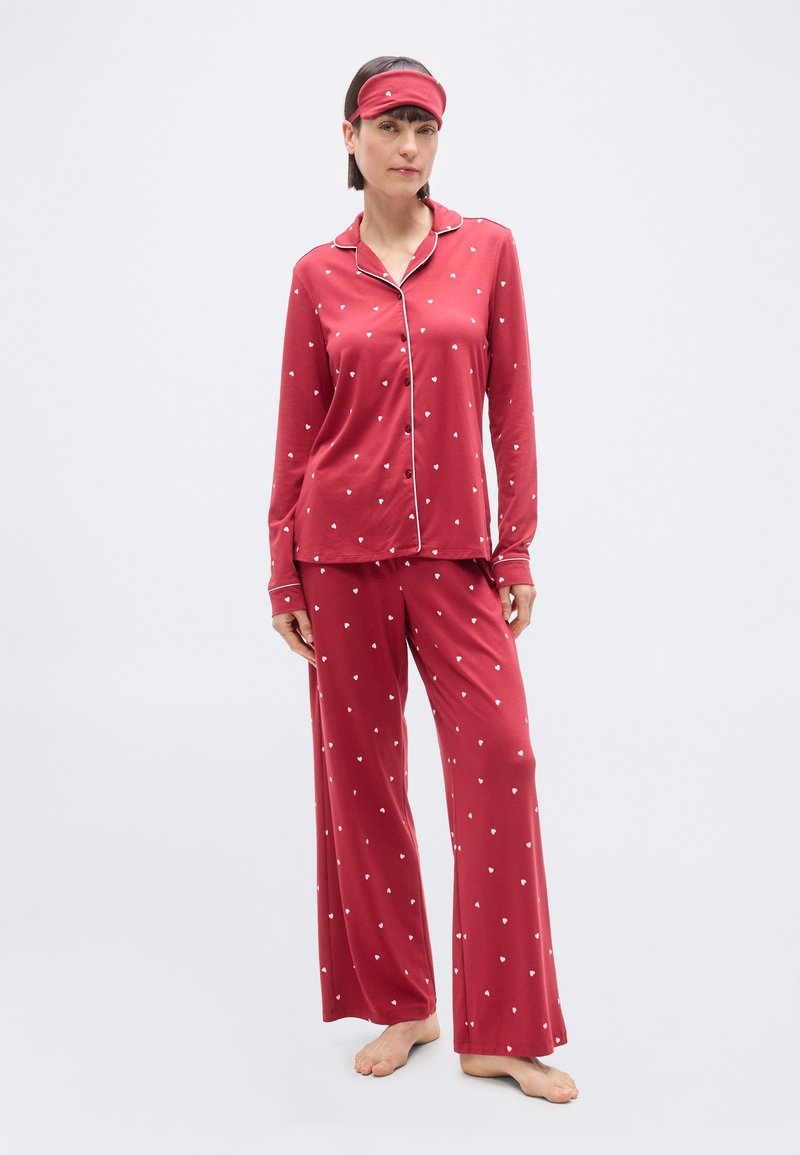 Femme debout, pieds nus, portant un pyjama boutonné à manches longues avec des motifs de cœurs rouges, un pantalon assorti et un masque de sommeil sur le front.