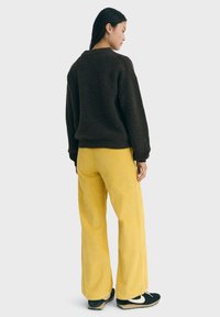 Donna che indossa un maglione di maglia marrone scuro, pantaloni a gamba larga di velluto a coste gialli e sneakers nere con il logo bianco, in piedi di spalle.