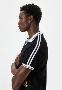 Svart polo t-shirt med vita detaljer och ränder på ärmarna, med en texturerad tyg och klassisk krage design. Modellen poserar i profil.
