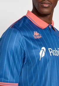 Mėlynas dryžuotas sportinis polo marškinėlis su rožine apykakle. Ant marškinėlio yra oranžinis Kappa logotipas ir baltas rėmėjo logotipas. Lygios tekstūros.