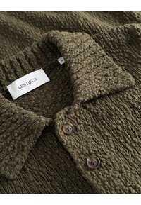 Olivgrön texturerad stickad skjorta med krage, utrustad med två bruna knappar och en etikett som säger "LES DEUX," storlek medium.