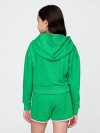 Dievča otočené chrbtom, oblečené v zelenej mikine a zodpovedajúcich zelených šortkách s ružovým lemovaním na pozadí s jednoduchým bielym pozadím.