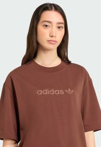 Camiseta extragrande marrón de algodón. Presenta un cuello acanalado y un logo de Adidas en relieve en la parte frontal. Textura suave, mangas cortas.