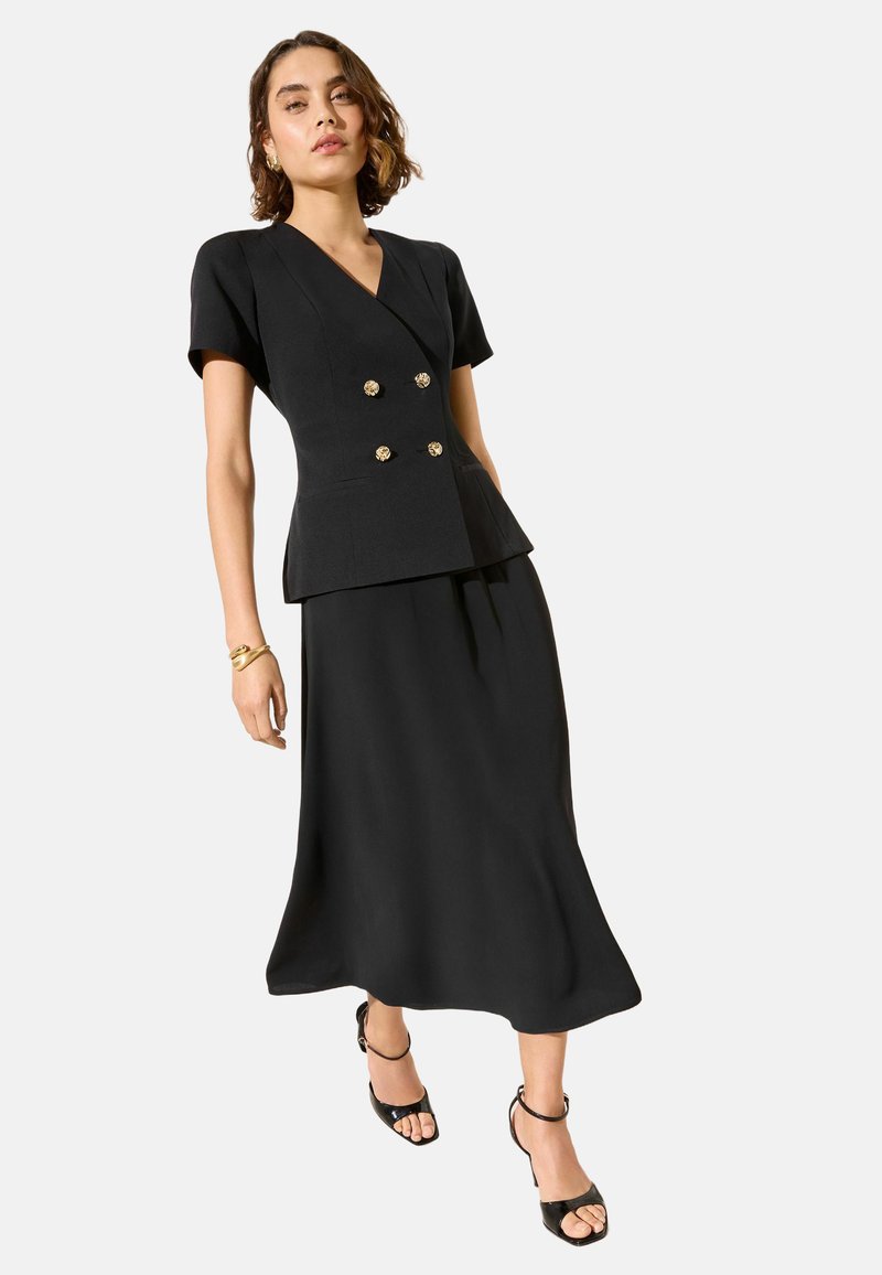 Frau präsentiert einen schwarzen Blazer mit kurzen Ärmeln und goldenen Knöpfen sowie einen passenden Midi-Rock, dazu schwarze Sandalen mit Absatz und ein goldenes Armband.