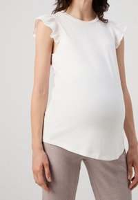 Top sin mangas blanco con acentos de encaje rizado en los hombros; tejido suave y liso; diseño ajustado alrededor de un vientre redondeado; combinado con pantalones beige acanalados.