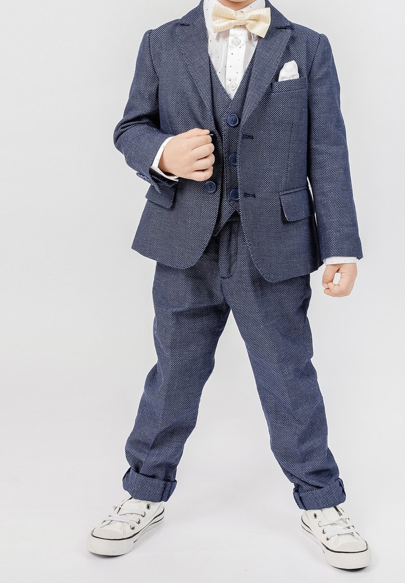Traje azul marino con un patrón texturizado, que incluye un blazer de tres botones, pantalones a juego, camisa blanca y pajarita color crema, combinado con zapatillas.