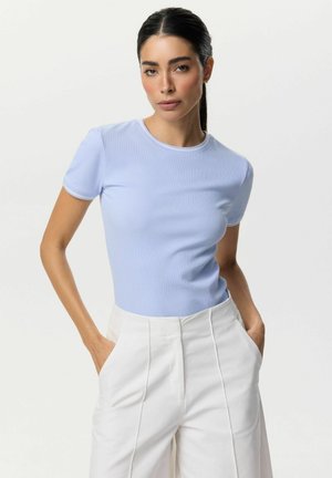 Kobieta z ciemnymi włosami związanymi w kucyk, ubrana w jasnoniebieski prążkowany krótki top i białe spodnie z wysokim stanem, z rękami w kieszeniach.
