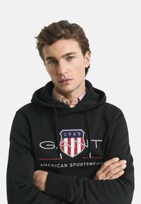 Felpa nera con cappuccio e logo "GANT" ricamato in bianco e rosso con design a scudo, laccetti, e una texture morbida. Indossata sopra una camicia con colletto.