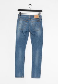 Blå denimjeans med bleknad tvätt hängande på svart galge mot vit bakgrund, som visar bakfickor och lädermärkeslapp.