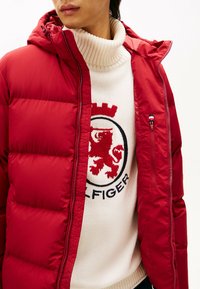 Rød puffjakke med hætte, featuring en lynlås lukning, quiltet design og sidelommer. Bærer den over en cremefarvet turtleneck sweater med grafisk detalje.