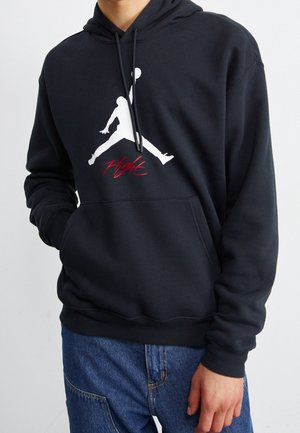 Person trägt schwarzen Kapuzenpullover mit weißem Basketballspieler-Logo und rotem Schriftzug "Flight", kombiniert mit blauen Jeans.