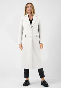 Long manteau blanc avec un col, deux boutons et des poches latérales. Porté par-dessus un haut noir et associé à un pantalon noir et des mocassins.