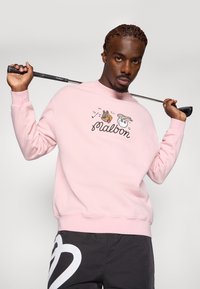 Felpa rosa chiaro con grafiche ricamate di un cane e una pallina da golf, scollo rotondo, polsini a coste, indossata con pantaloni neri con un logo a forma di cuore.