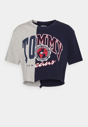 T-shirt court bicolore, moitié gris, moitié bleu marine, divisé verticalement avec le logo "Tommy Jeans" et un emblème rouge, blanc et noir centré.
