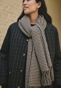 Beige and navy check-patterned scarf made of soft material, featuring fringed edges, worn over a dark plaid coat with gold hardware.