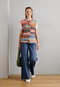 Strickweste mit mehrfarbigem Streifenmuster, mit Knöpfen vorne. Kombiniert mit weit geschnittenen blauen Jeans und weißen Turnschuhen.