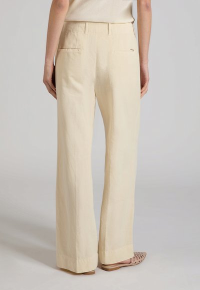 Mujer con pantalones anchos de talle alto en color beige y zapatos planos tejidos beige, vista desde atrás sobre un fondo liso.