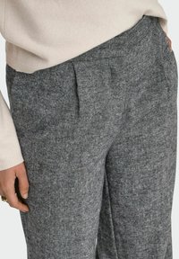 Kairėje kelnių kišenėje, aptakiose pilkai tekstūruotose kelnių, užsitempusiose virš liemens, matoma nuo liemens iki vidurio šlaunies, su kremu ilgesniais rankovėmis.