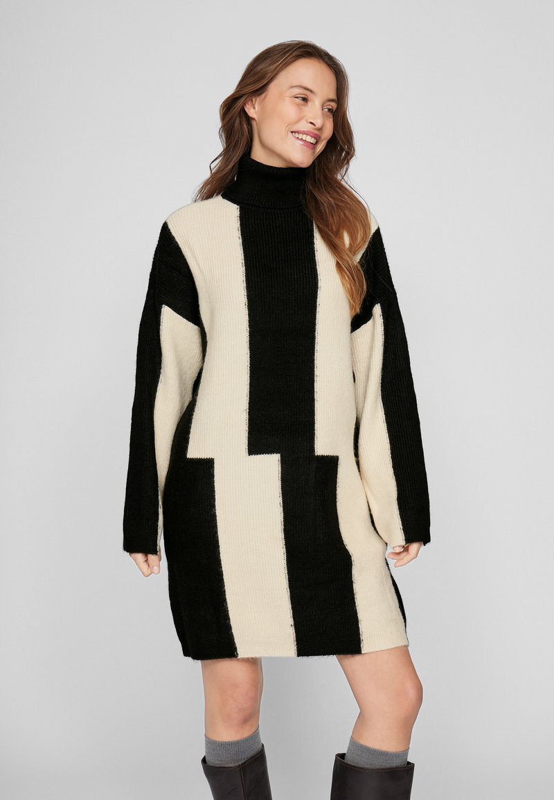 Lielizmēra turtlenecka kleita melnā un krēmkrāsā, ar ģeometriskiem rakstiem un ribotu tekstūru. Garās piedurknes sniedzas līdz plaukstām.