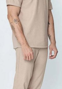 Haut à manches courtes en tricot côtelé beige clair associé à un pantalon assorti. Le matériau semble doux avec une coupe décontractée ; tatouages visibles sur le bras.