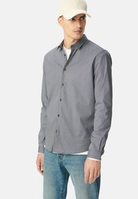 Camicia a maniche lunghe grigia con un motivo geometrico, taschino sul petto, chiusura con bottoni e polsini a contrasto, abbinata a jeans azzurri chiari.