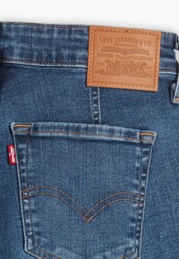 Dark blue denim jeans feature a leather patch with embossed text and a red tab on the rear pocket. The stitching is in a contrasting orange color.